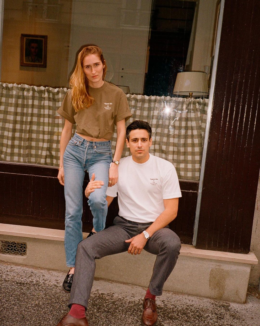A young couple poses in front of a shop window, the woman stands while the man sits, both wearing casual outfits and a neutral expression.