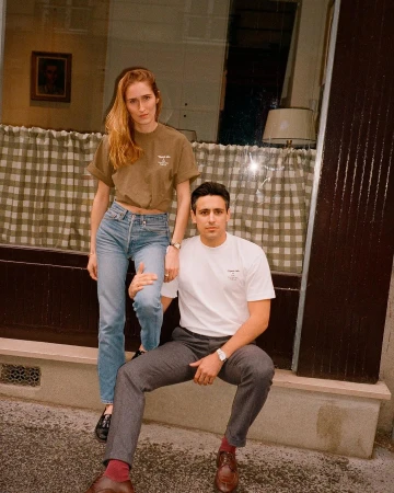 A young couple poses in front of a shop window, the woman stands while the man sits, both wearing casual outfits and a neutral expression.