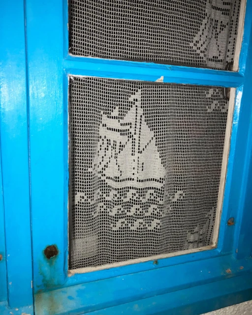 Close-up of a vintage blue window with a white, crocheted curtain featuring a ship design, adding a nautical touch to the interior.