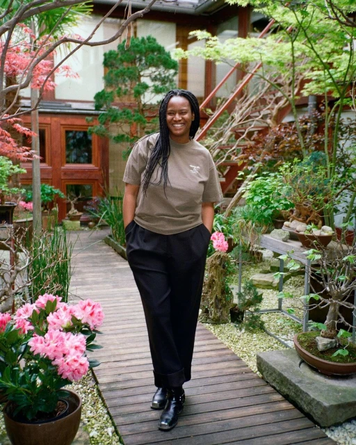 A smiling Black woman strolls a garden path lined with flowering bushes and bonsai trees in front of a Japanese-style building with stairs.