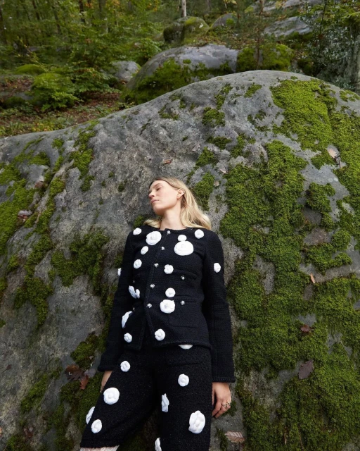 Adult female model in a black and white rose-embellished jacket and pants poses near a mossy rock formation in an outdoor wooded area.