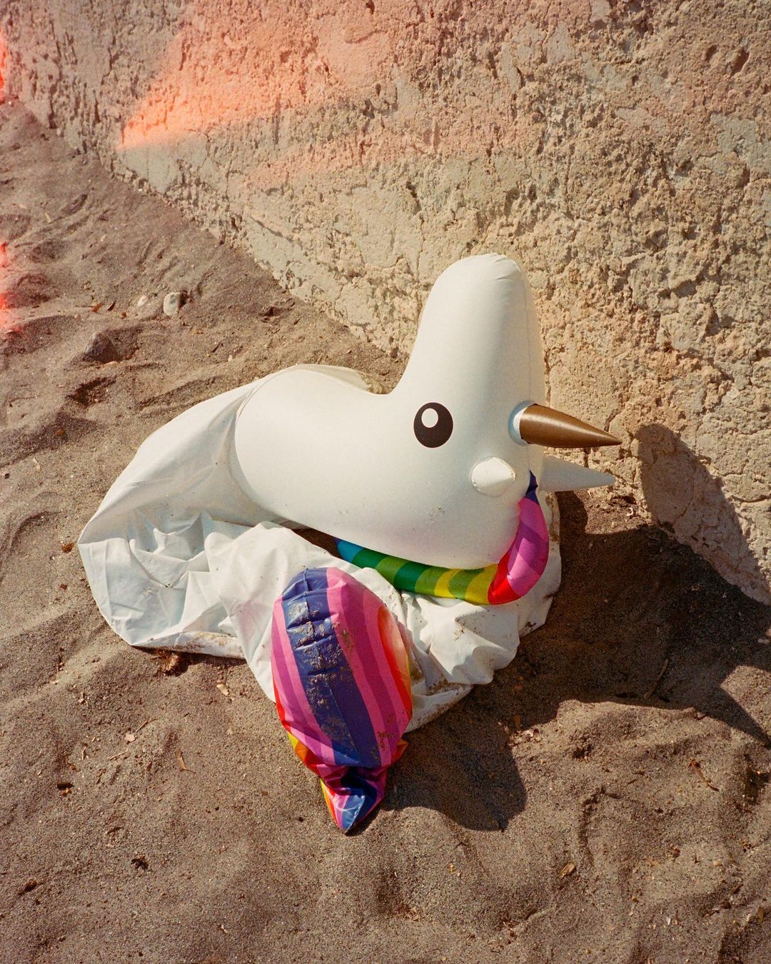 Inflatable unicorn on a sandy surface near a textured wall. The unicorn has a rainbow mane and tail, with soft, diffused lighting.