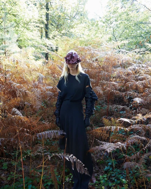 A slender woman in a long black dress and ornate hat stands in a dense forest. Dried bracken surrounds her in autumnal light.