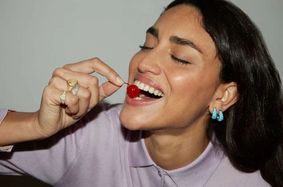 Portrait of a smiling brunette woman with fair skin holding a cherry in her mouth, wearing gold rings and blue hoop earrings in an indoor setting.