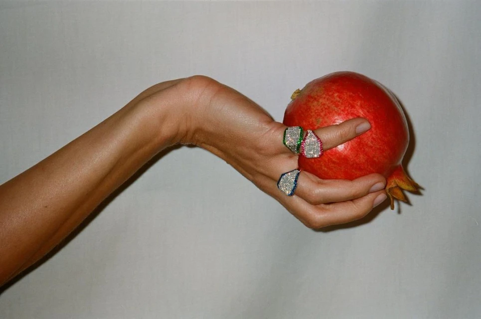 Close-up of a fair-skinned woman's hand holding a ripe pomegranate, adorned with three sparkling geometric rings. 