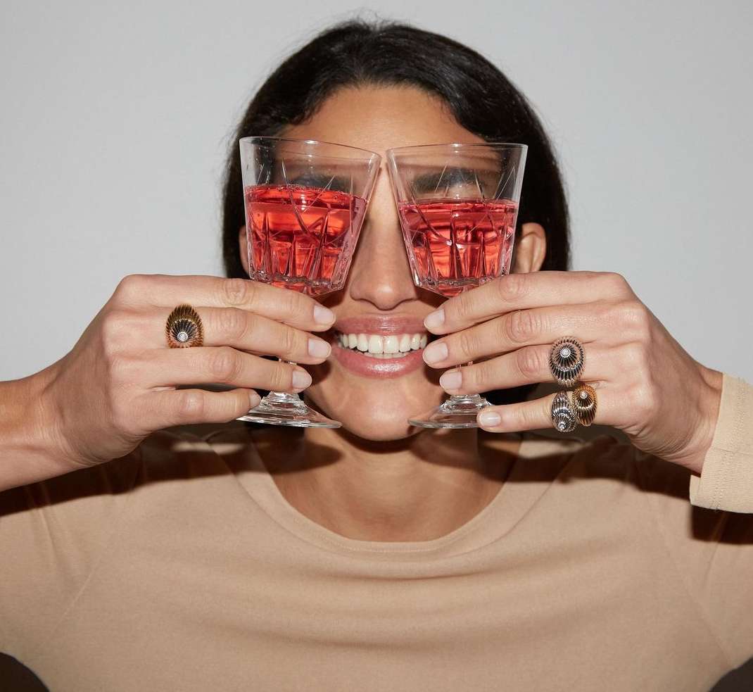 A cheerful woman smiles while holding crystal glasses filled with rose-colored liquid, wearing rings, against a neutral backdrop.