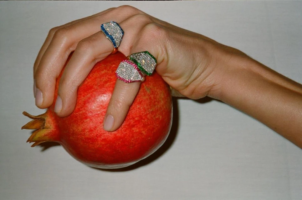 Close-up of a woman's hand with three colorful rings holding a ripe pomegranate against a white backdrop.
