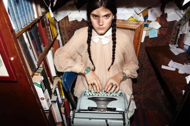Woman with braided dark hair wearing a beige blouse typing on a vintage typewriter in a cluttered book-filled room
