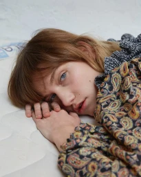 Close-up portrait of a woman with red hair and blue eyes resting her head on her hands, wearing a patterned paisley top.