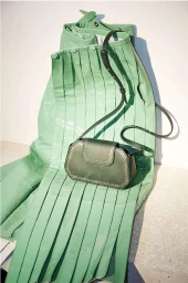 Green leather shoulder bag resting on a glossy pastel green pleated garment against a white textured wall.
