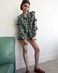 Model standing against a white wall wearing a green plaid ruffled tunic and brown leather clogs, photographed by Thierry Lebraly.