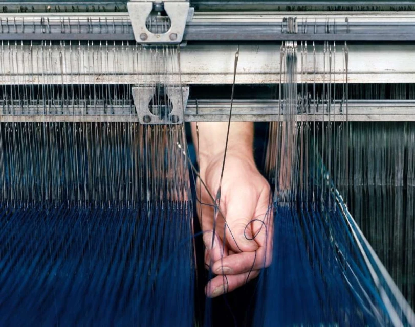 Close-up of a weaving machine with a person's hand guiding blue thread. Industrial loom in operation, textile production process.