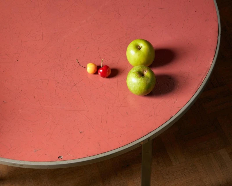 Overhead shot of two green apples and two cherries (one yellow, one red) arranged on a round, textured, coral-colored table top with silver trim.