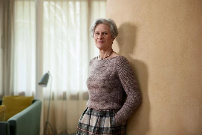 Elderly woman with grey hair wearing a patterned sweater leaning against a textured wall in a bright interior room