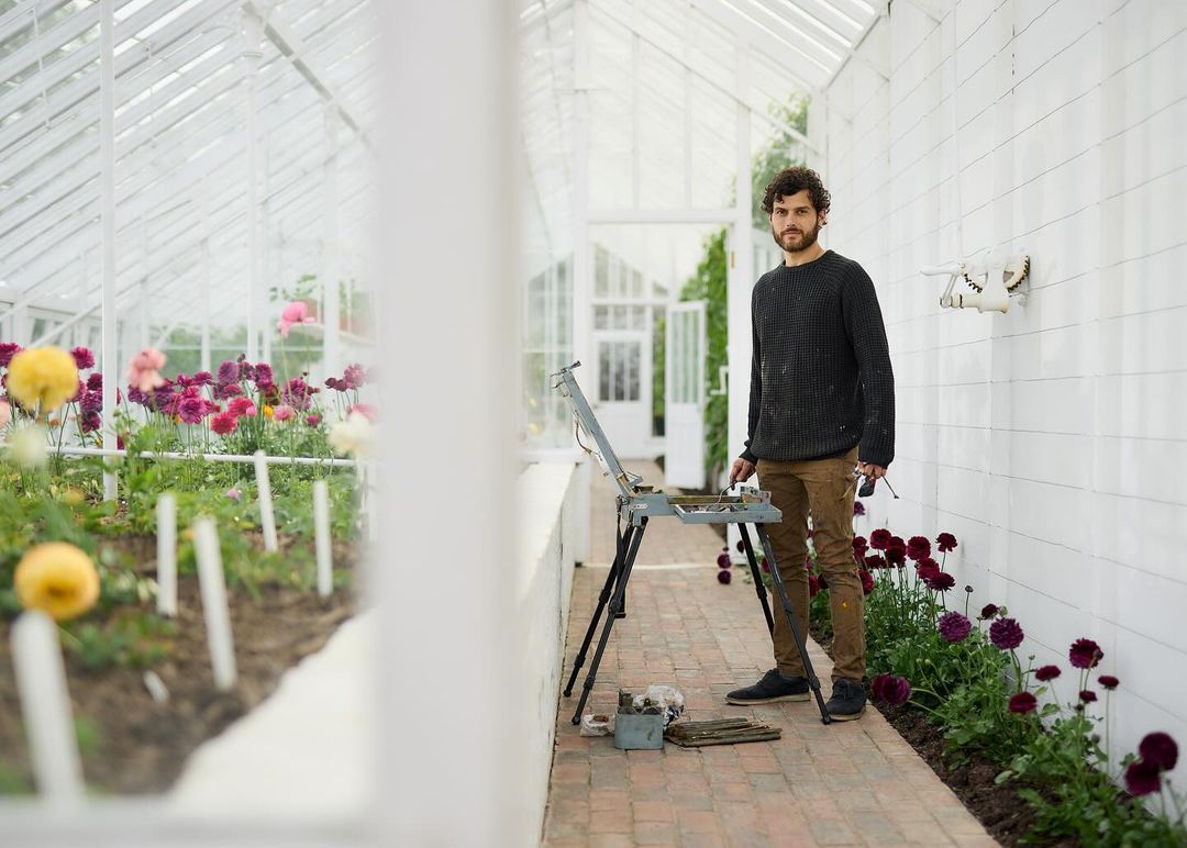 Adult male artist stands in a greenhouse surrounded by blooming flowers and an easel, dressed in casual clothing in natural light.