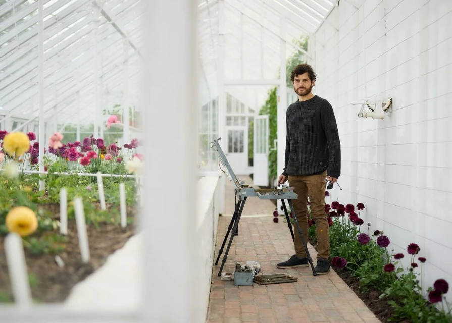 Adult male artist stands in a greenhouse surrounded by blooming flowers and an easel, dressed in casual clothing in natural light.