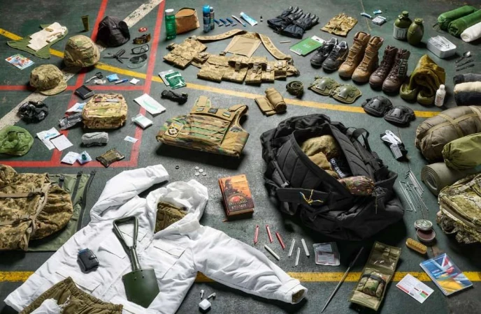 Overhead studio shot displays arranged military gear including camouflage uniforms, boots, packs, helmets, and tools laid out on a gray floor.