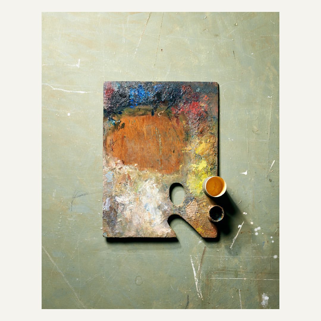 Overhead studio shot of an artist's wooden palette covered in colorful paint smears next to two paint jars on a muted green surface.