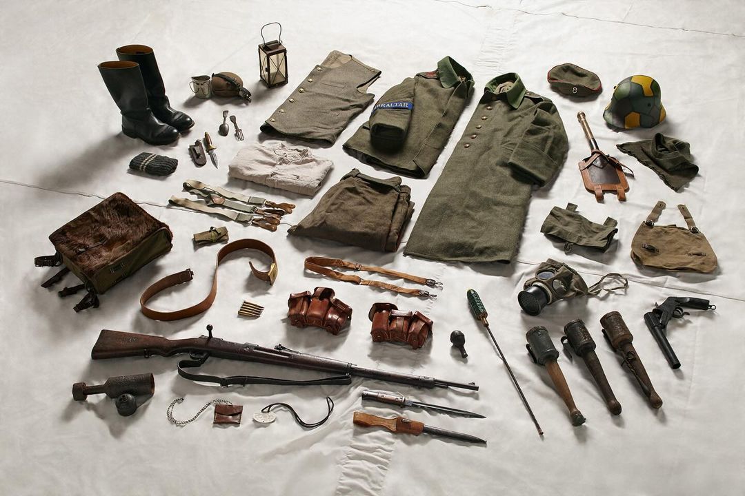 Arrangement of vintage military gear including uniforms, boots, bags, weapons, and accessories laid out on a white surface, flat lay.