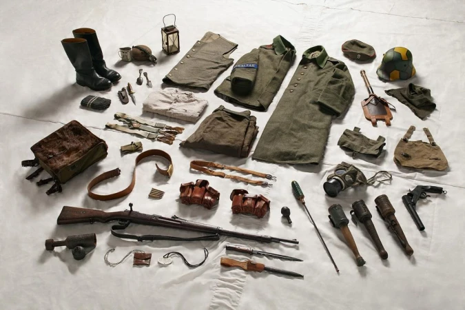 Arrangement of vintage military gear including uniforms, boots, bags, weapons, and accessories laid out on a white surface, flat lay.