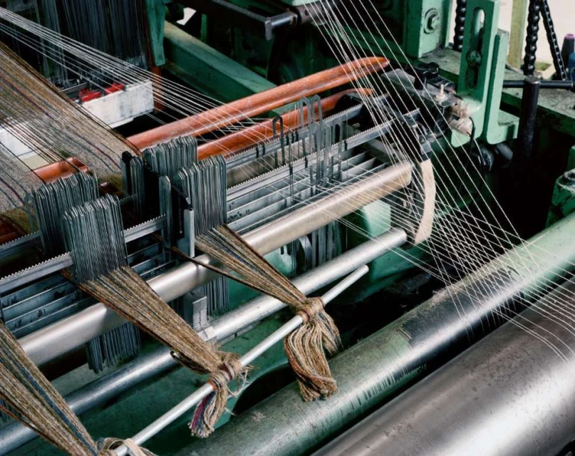 Close-up of a weaving machine with visible threads and metallic components in industrial setting, highlighting textile production.