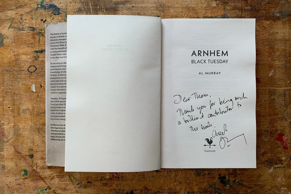 Close-up shot of an open book on a wooden table featuring the title 'Arnhem Black Tuesday' and a handwritten note and signature.