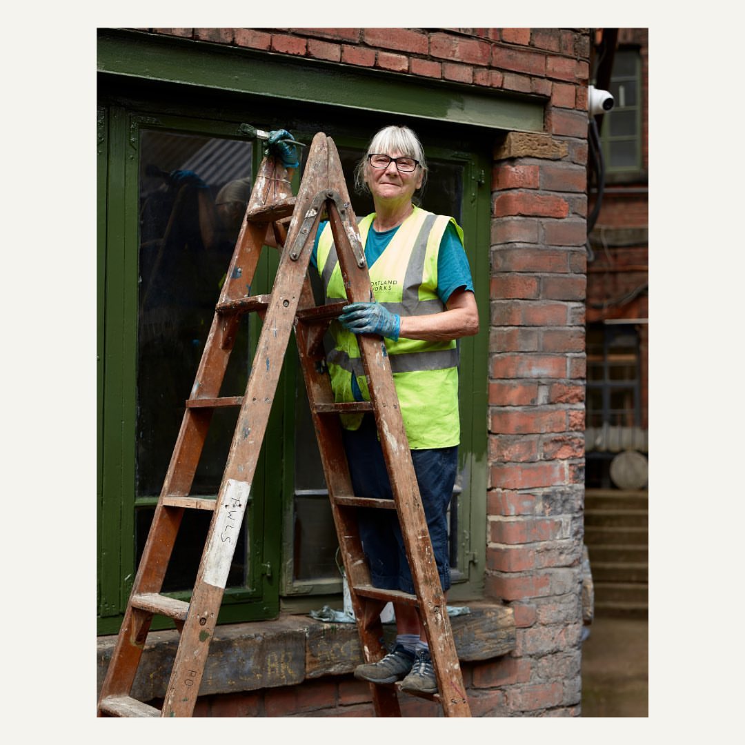 Elderly woman stands on ladder outdoors wearing safety vest, gloves, painting window frame green. Brick wall, daylight, trees in background.