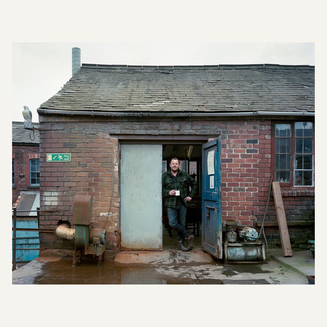 A man leans in doorway of a brick workshop, holding a cup. Vintage industrial exterior. Brick building with green door.