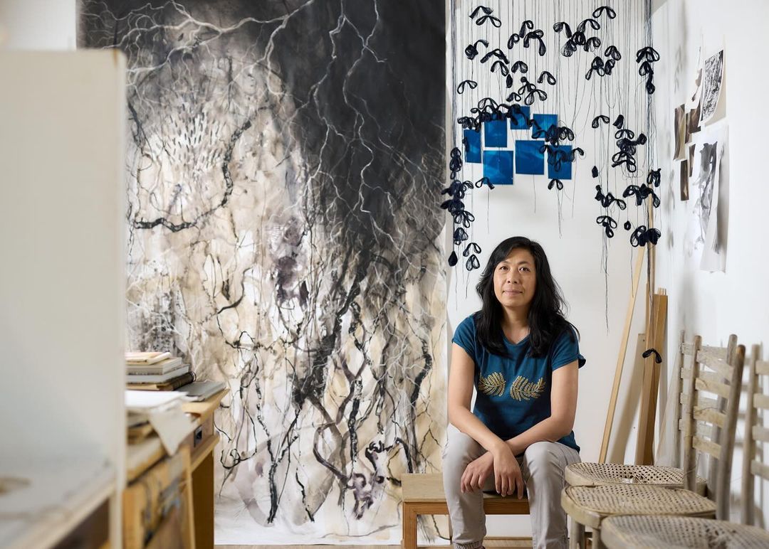 Asian woman sitting in an art studio in front of large-scale abstract drawing and installation hanging from the ceiling.