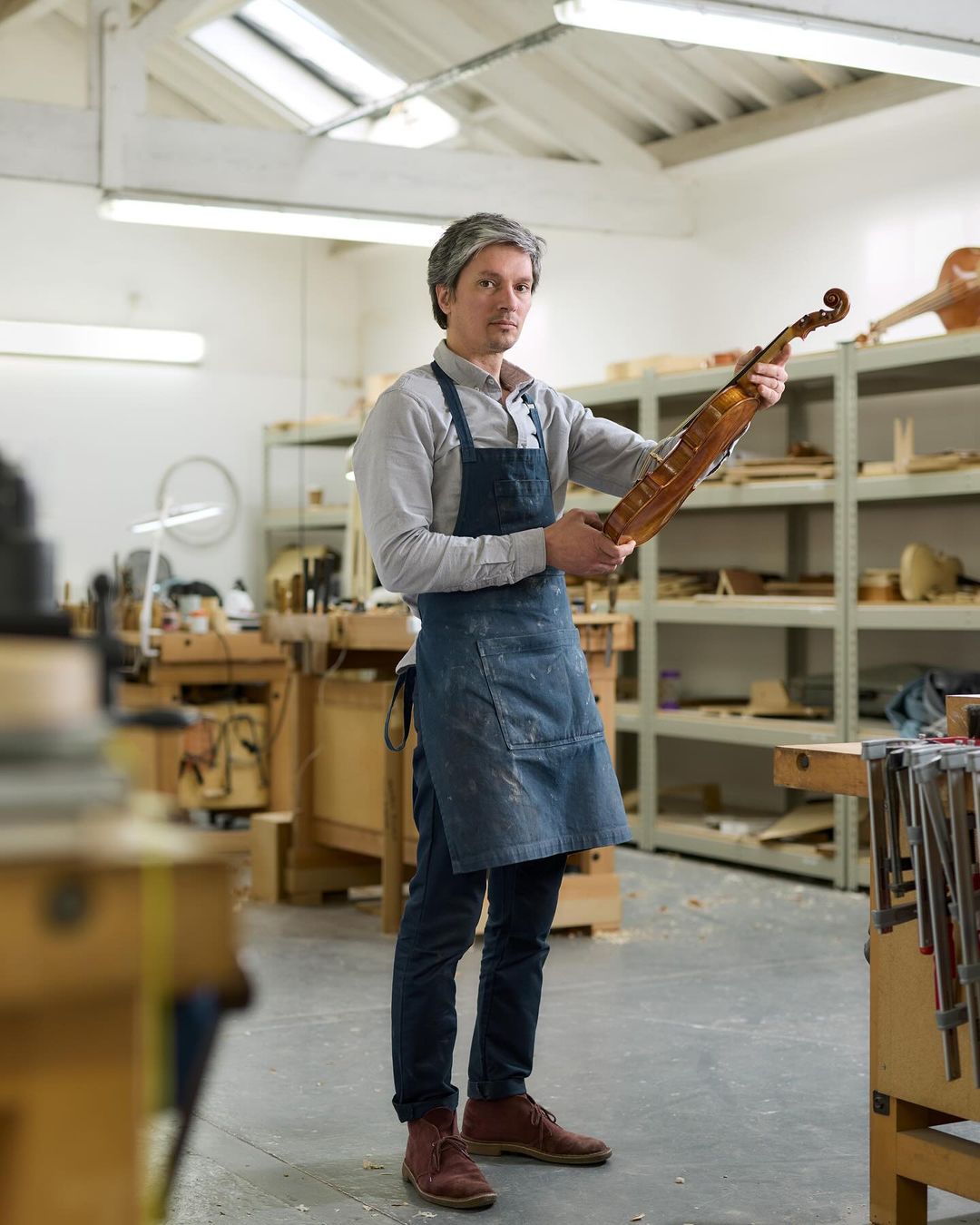Man in a workshop holding a violin, wearing an apron. Interior studio with workbenches, storage, and tools.