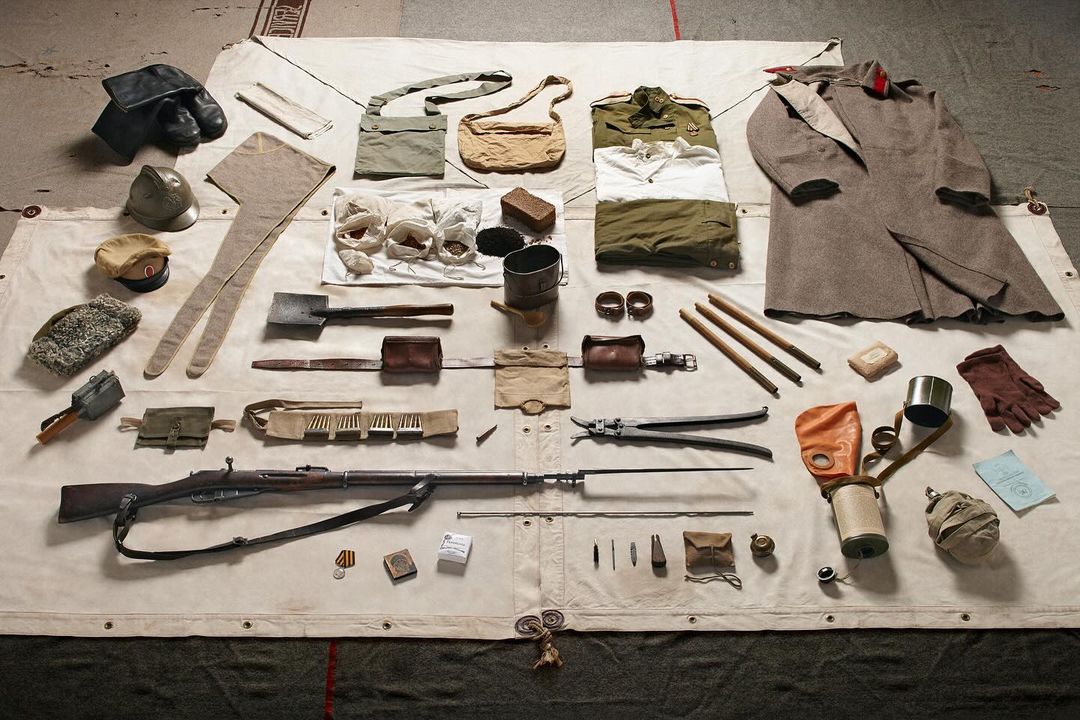 Overhead shot of vintage military gear including a rifle, clothing, helmets, bags, tools, and protective gear, displayed on a neutral surface.