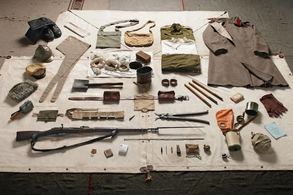 Overhead shot of vintage military gear including a rifle, clothing, helmets, bags, tools, and protective gear, displayed on a neutral surface.