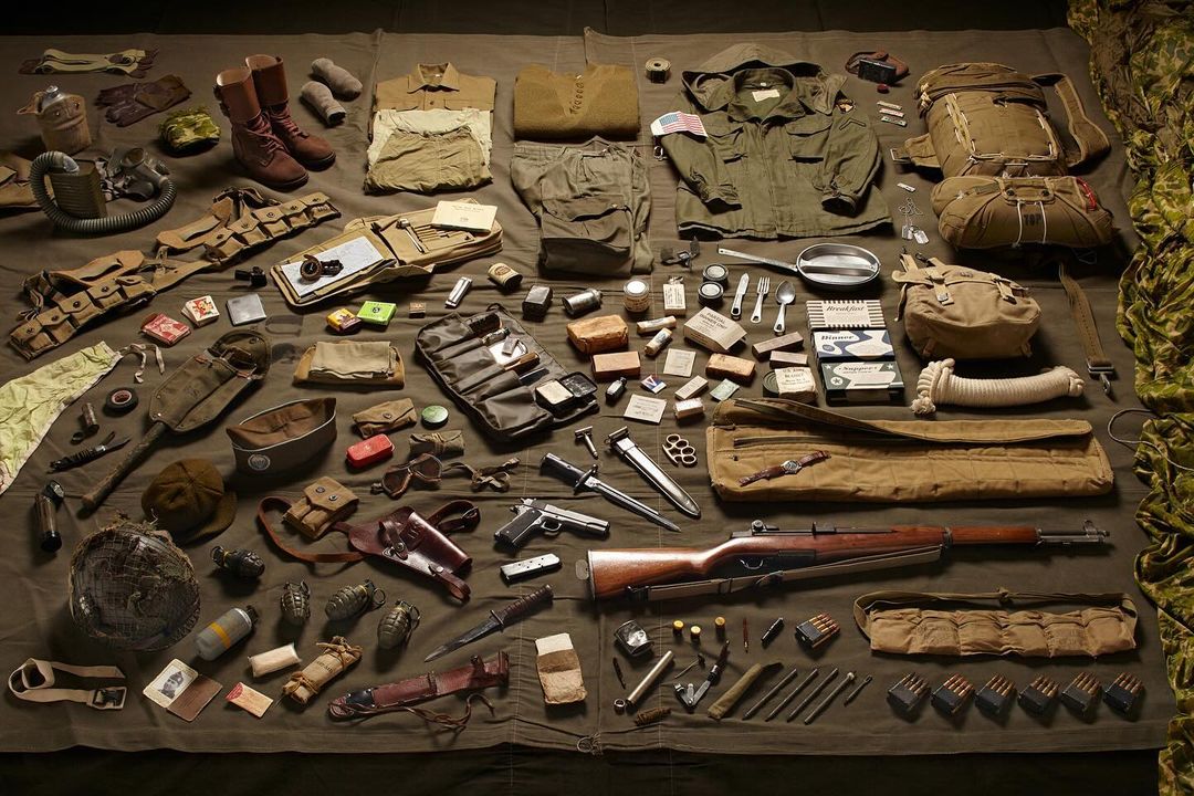 Collection of vintage WWII-era military equipment, including uniforms, weapons, boots, helmets, and tactical gear, laid out on a khaki-colored cloth.