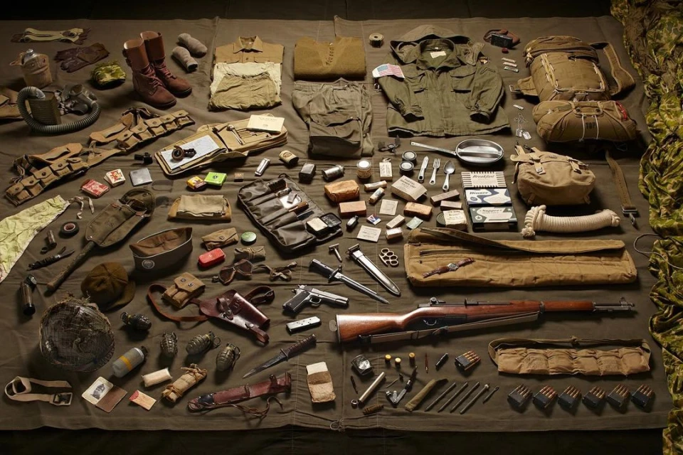 Collection of vintage WWII-era military equipment, including uniforms, weapons, boots, helmets, and tactical gear, laid out on a khaki-colored cloth.