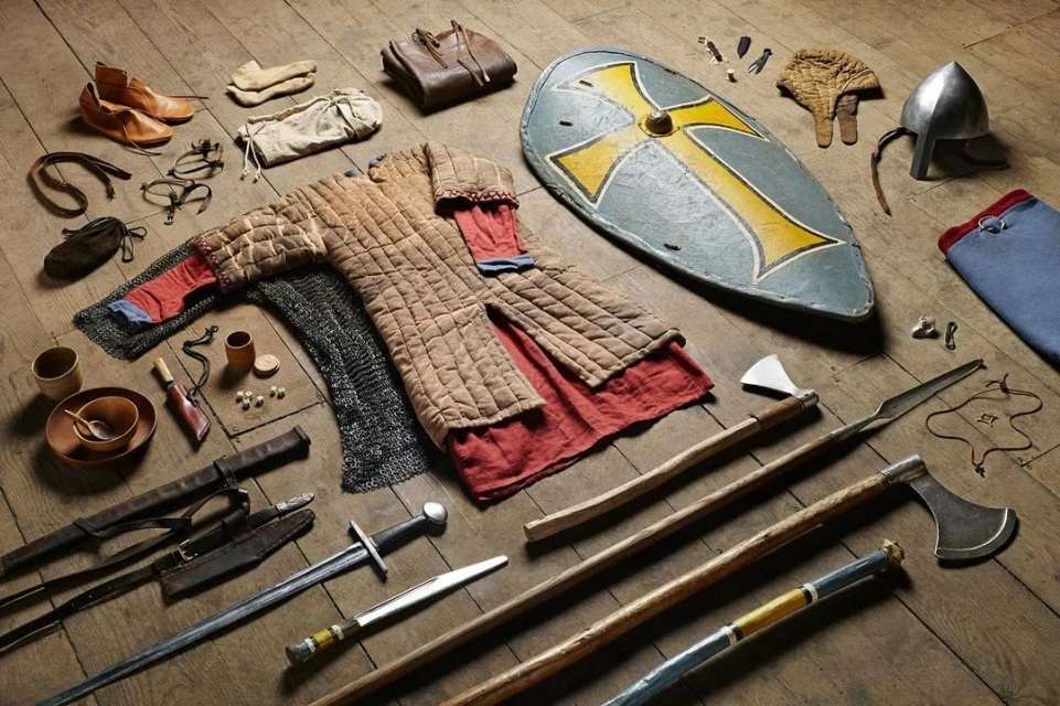Still life featuring replica medieval-era clothing, footwear, weaponry, armor, and accessories arranged on a light brown wooden surface.