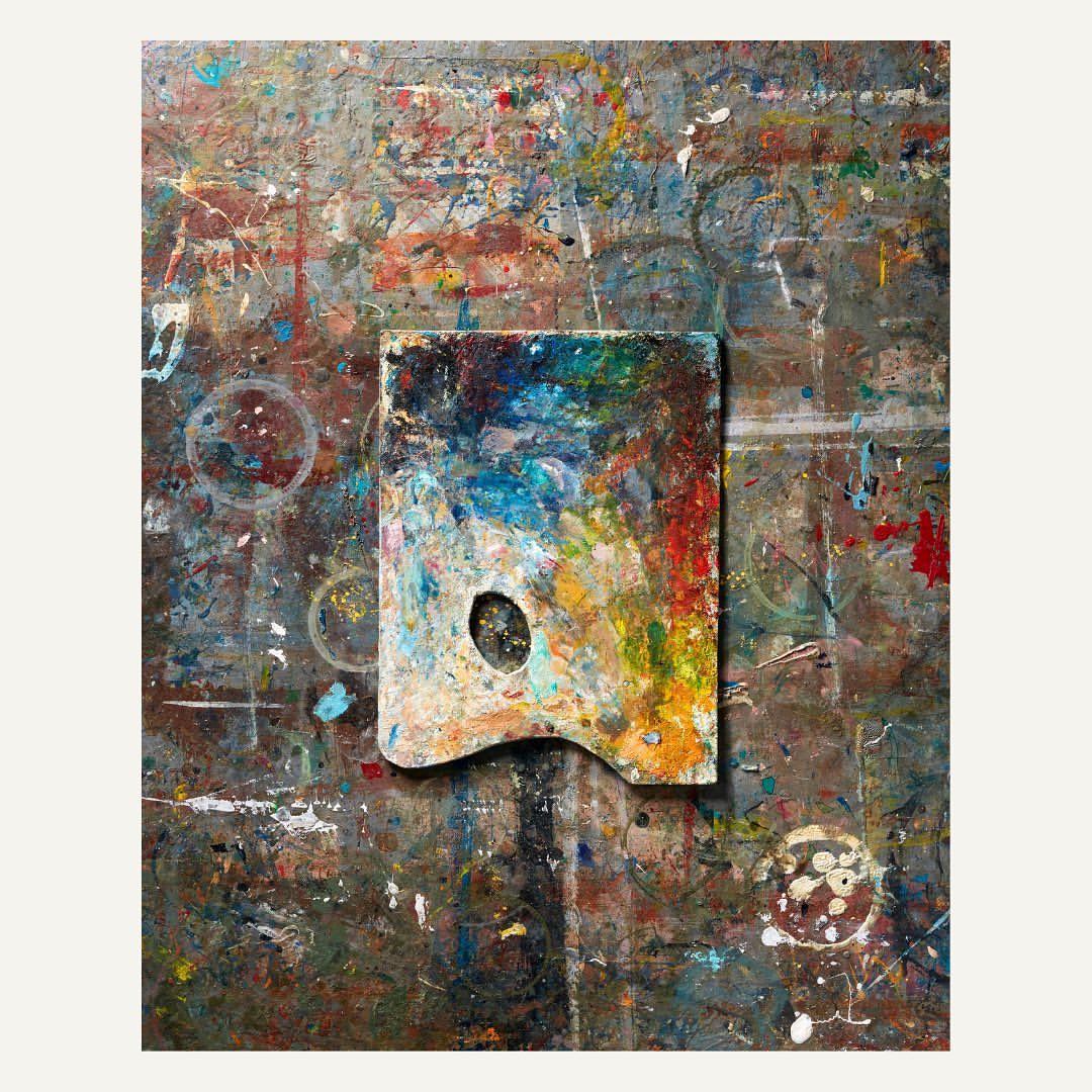 Overhead studio shot of a colourful paint palette on a splattered and textured grey surface, suggesting artistic process.
