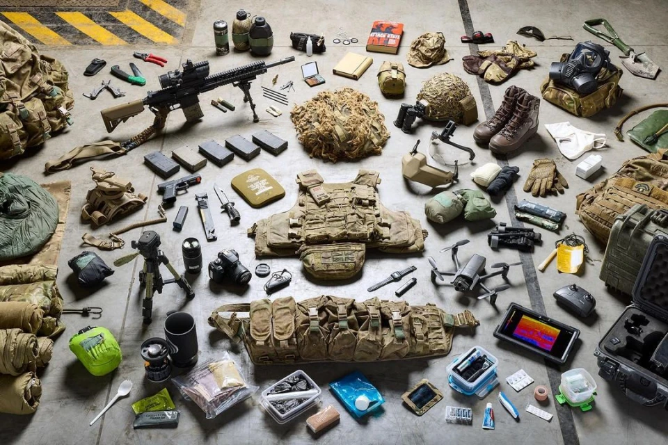 Overhead studio shot of tactical military gear, including a rifle, vest, drone, helmet, and food supplies, arranged on a gray surface.