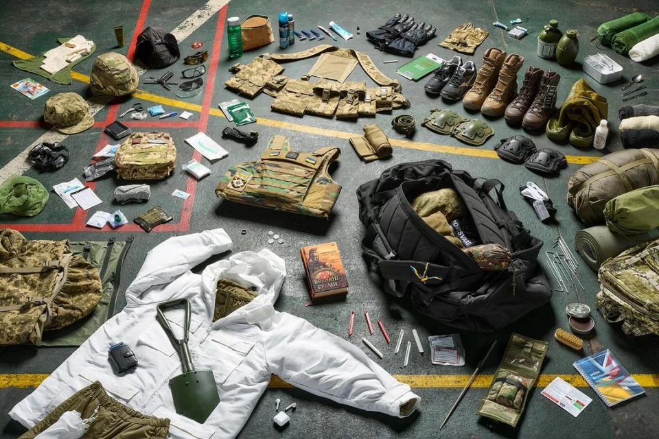Overhead shot of military gear including boots, camouflage clothing, vests, packs, shovels, tents, and supplies laid out on a marked floor.