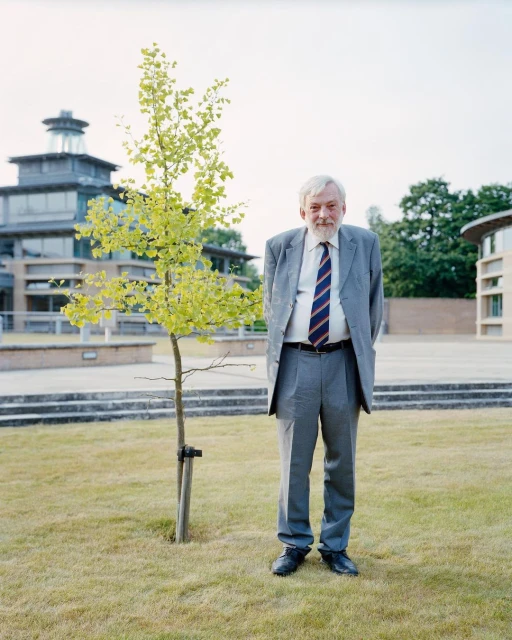 Full-length portrait of an adult man in a gray suit and tie standing on a lawn with a tree, with buildings in the background.