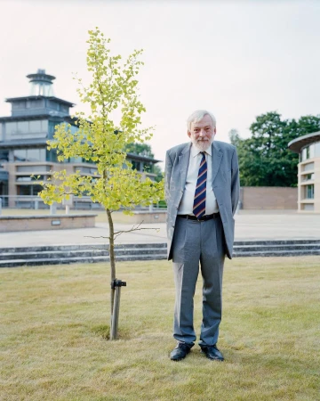 Full-length portrait of an adult man in a gray suit and tie standing on a lawn with a tree, with buildings in the background.