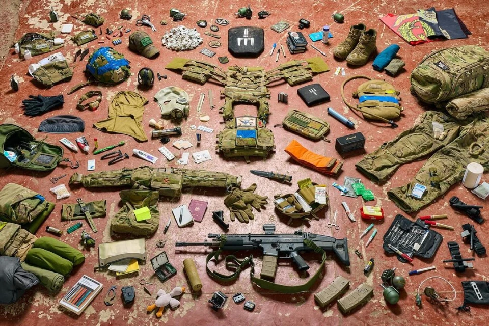 Still life of tactical military gear laid out on a reddish-brown surface. Camouflage clothing, rifle, and other equipment are displayed.