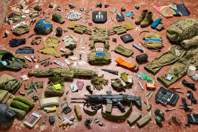 Still life of tactical military gear laid out on a reddish-brown surface. Camouflage clothing, rifle, and other equipment are displayed.