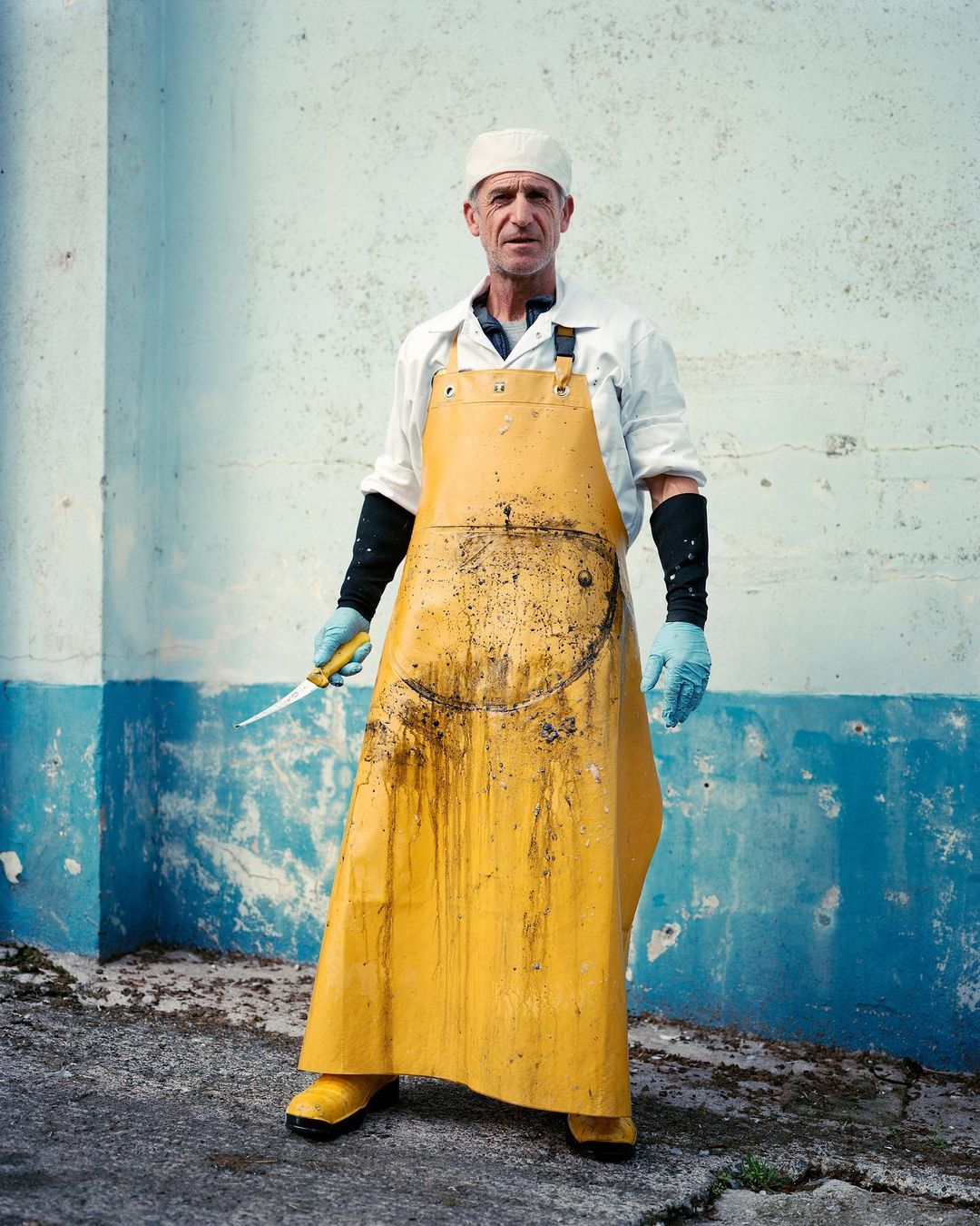 Full shot of a serious-looking man in a yellow apron, cap, and gloves holding a knife in an indoor setting with a textured wall.