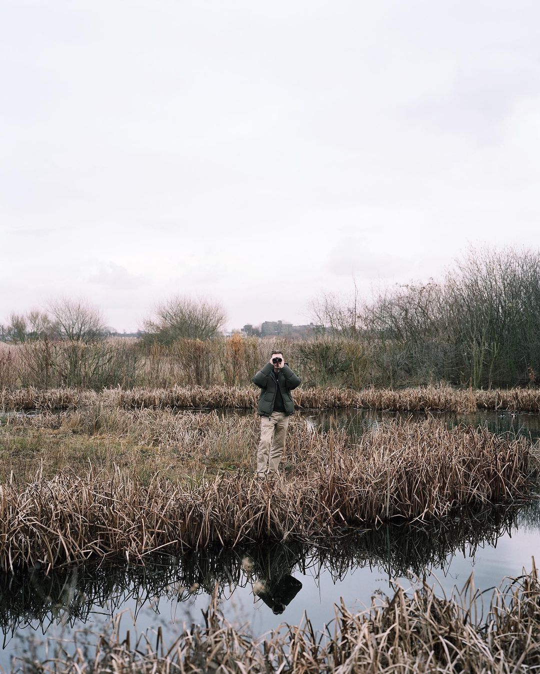 Adult stands in marshland water, holding binoculars. Grassy reeds surround the figure, under a cloudy sky, with a reflection in the water.