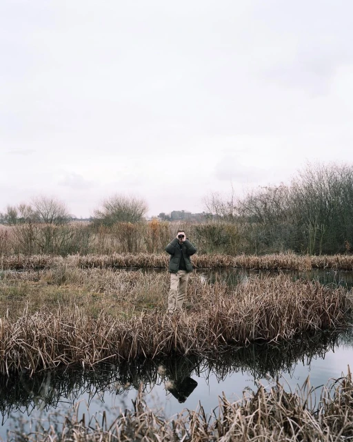 Adult stands in marshland water, holding binoculars. Grassy reeds surround the figure, under a cloudy sky, with a reflection in the water.