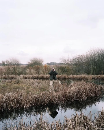 Adult stands in marshland water, holding binoculars. Grassy reeds surround the figure, under a cloudy sky, with a reflection in the water.