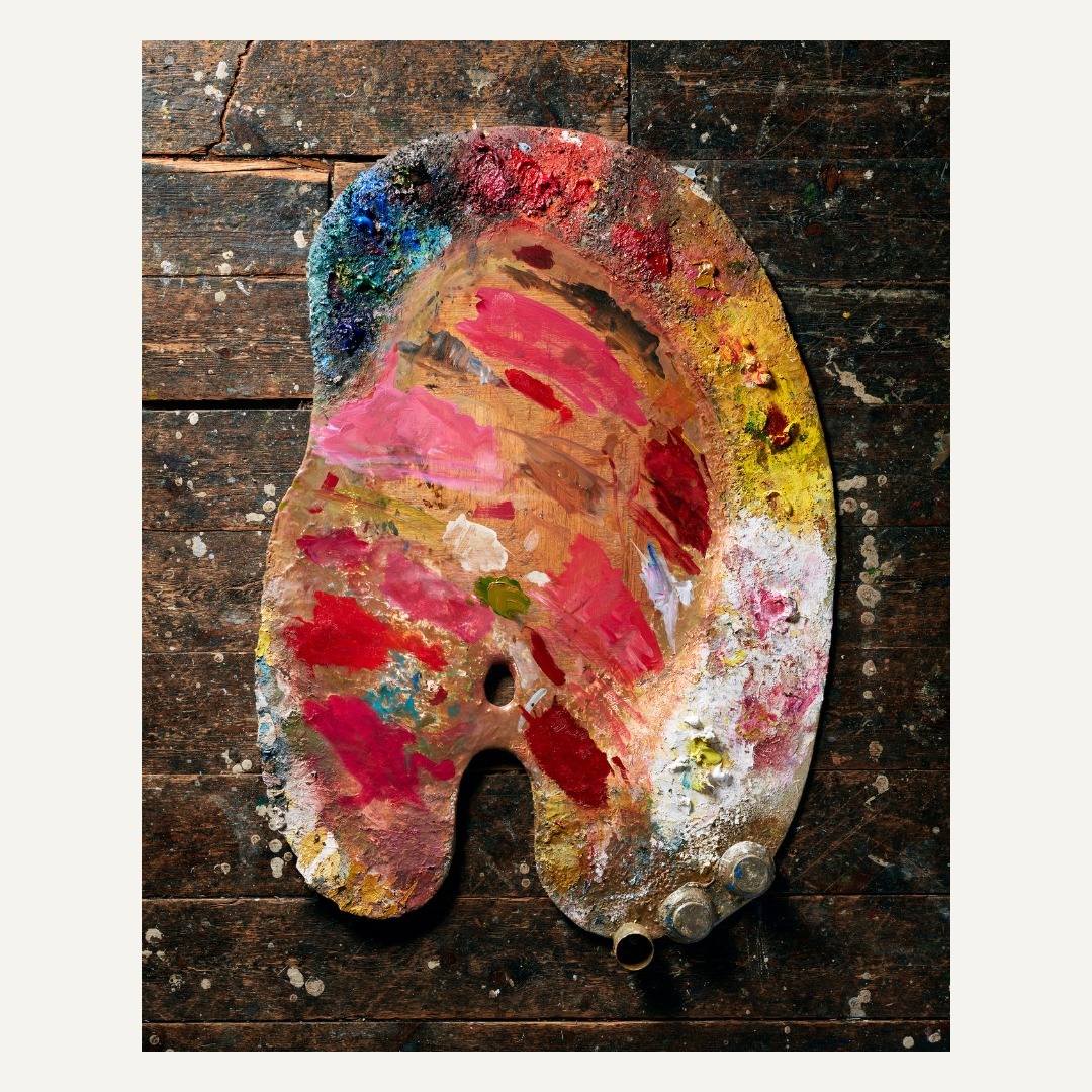 Overhead shot of a colorful paint palette filled with red, yellow, blue, and pink hues, sitting on a dark brown wooden surface.