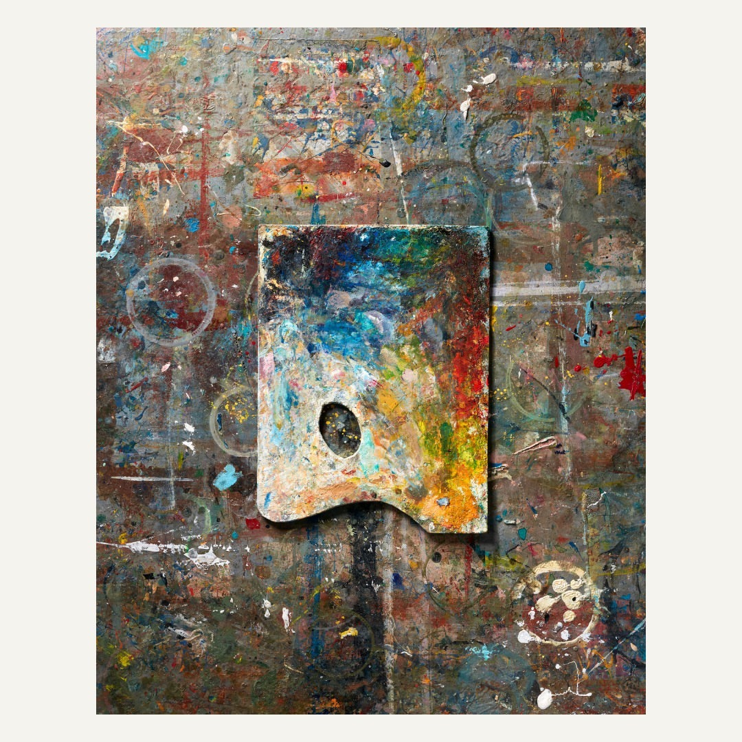 Close-up shot of a paint-covered wooden palette on a paint-splattered surface, showcasing a colorful array of paints and textures.