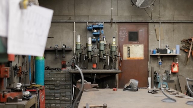 Industrial workshop interior featuring metalworking machinery, work tables, and a cluttered yet functional workspace, evoking a utilitarian atmosphere.