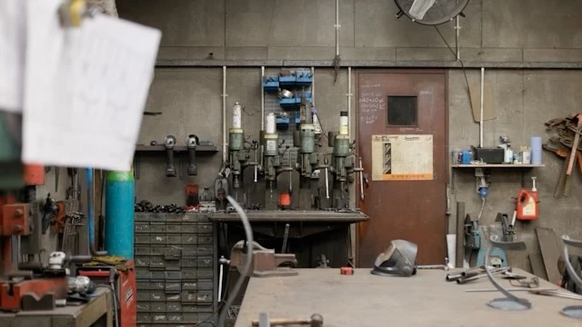 Industrial workshop interior featuring metalworking machinery, work tables, and a cluttered yet functional workspace, evoking a utilitarian atmosphere.